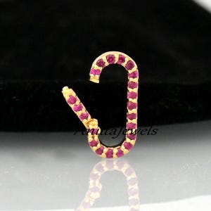 Puede incluir: Un amuleto dorado con un diseño incrustado de piedras rosas en forma de gancho. El amuleto es pequeño y delicado.