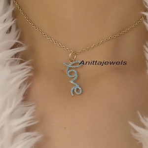 Pode incluir: Um colar de corrente de ouro com um pingente que diz "LOVE" em letras cursivas. As letras são contornadas por pequenas pedras azuis.