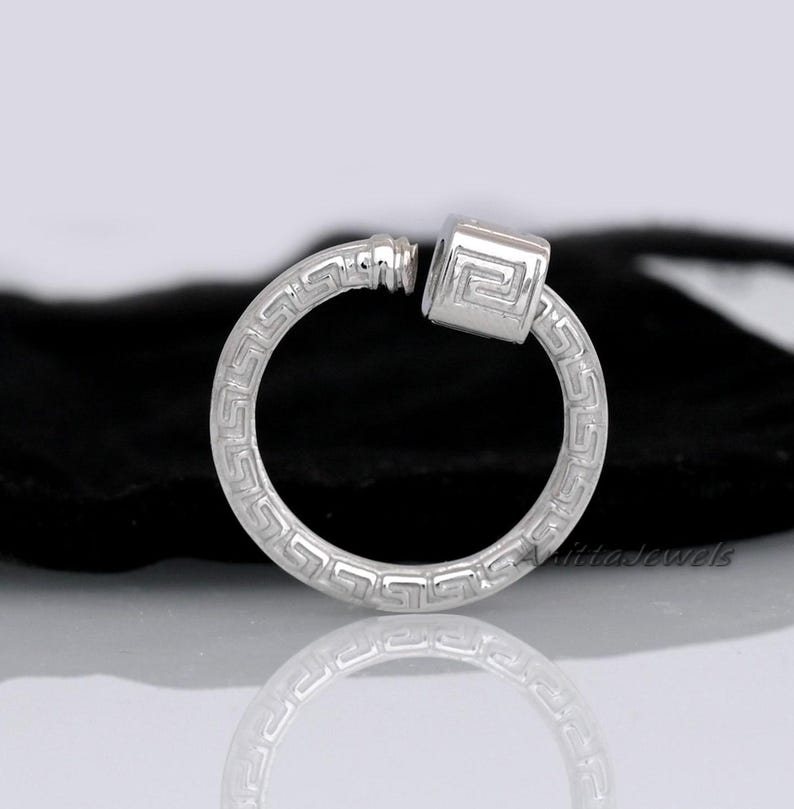 May include: A silver-colored hoop earring with a Greek key pattern. The earring features a square-shaped charm with a matching design. The earring is shown against a dark background.
