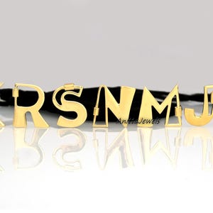 Pode incluir: Letras de metal dourado que soletram a palavra "KRSNMJT" em uma fita preta. As letras são todas maiúsculas e têm uma fonte ligeiramente arredondada.