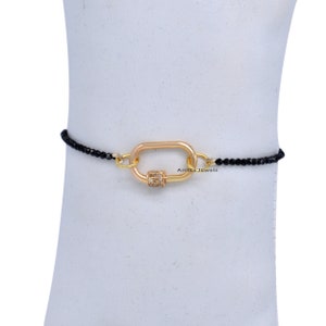 Puede incluir: Un brazalete de cuentas negras con un cierre dorado con un diseño de diamantes pavé. El cierre tiene forma de rectángulo con esquinas redondeadas. El brazalete está en un maniquí blanco.