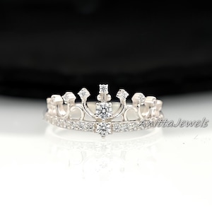 Puede incluir: Un anillo de corona plateada con un diseño de doble banda. La banda superior presenta una forma de corona con piedras preciosas transparentes. La banda inferior está forrada con piedras preciosas más pequeñas. Texto "Anitta Jewels".