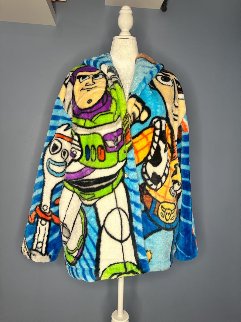 May include: A plush, hooded robe featuring colorful cartoon character designs. The robe showcases Buzz Lightyear, Woody, and other characters against a blue and white striped background. The robe is displayed on a mannequin.