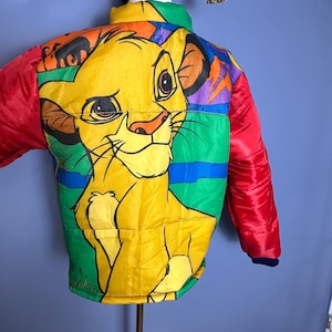 May include: A colorful, vintage puffer jacket featuring a large illustration of Simba from The Lion King. The jacket has a yellow Simba graphic on a green, blue, and orange background, with red sleeves and a navy blue collar.