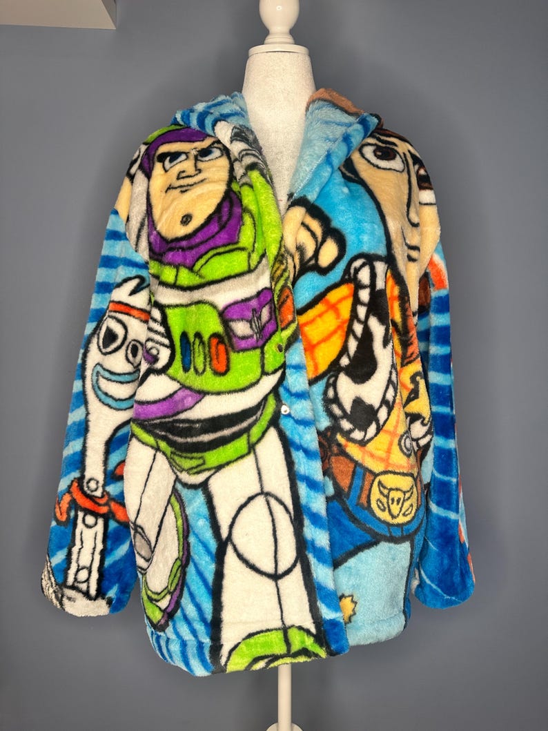 May include: A soft, hooded robe featuring characters from the animated film Toy Story. The robe displays Buzz Lightyear, Woody, and Forky in bright colours. The robe has a blue striped pattern on the sleeves and hood.