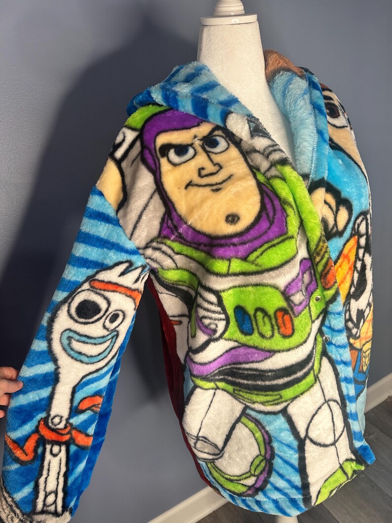 May include: A soft, hooded robe featuring characters from the animated film. The robe showcases Buzz Lightyear and Forky on a blue background with striped sleeves. The robe is made of soft, colourful fabric.