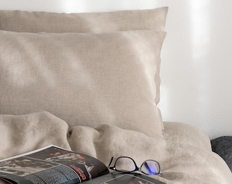 Puede incluir: Juego de cama de lino beige con dos fundas de almohada y una funda nórdica. Una revista abierta y gafas con montura negra descansan sobre la cama. La ropa de cama tiene un aspecto suave y texturizado, creando un ambiente acogedor.