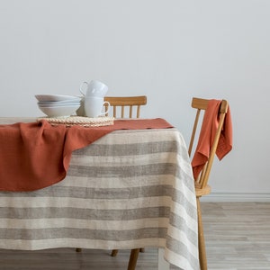 Könnte beinhalten: Eine gestreifte Tischdecke aus Leinen in Beige und Braun mit einem rostfarbenen Leinenläufer darauf. Weiße Keramikschalen und Tassen sind auf einem geflochtenen Platzdeckchen auf dem Tisch gestapelt. Zwei Holzstühle mit einer rostfarbenen Leinen-Serviette auf der Rückenlehne eines Stuhls.