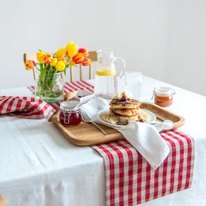 May include: A white tablecloth with a red and white checkered table runner. A wooden tray with a plate of pancakes, bananas, and a jar of jam. A glass of water and a pitcher of water are on the table. A bouquet of yellow and orange tulips is in a glass vase.