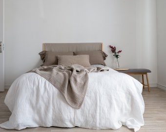 Puede incluir: Una funda nórdica de lino blanco con una manta de punto marrón encima. La cama tiene una cabecera de lino marrón y dos almohadas marrones. Una pequeña mesita de noche de madera con un jarrón de bayas rojas está a la derecha de la cama.