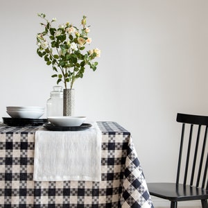 Pode incluir: Uma toalha de mesa xadrez preta e branca com um corredor de mesa de linho branco e um vaso de flores brancas sobre uma mesa. Uma cadeira de madeira preta está no fundo.