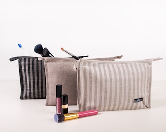May include: Three cosmetic bags in a row, each with a different striped pattern. The bags are filled with makeup brushes, a toothbrush, and lipsticks. One bag has black and gray stripes, the other two have beige and white stripes.