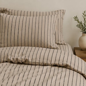 Könnte beinhalten: Beige Bettwäsche-Set mit schwarzen vertikalen Streifen. Das Set beinhaltet Kissen und einen Bettbezug. Die Streifen sind gleichmäßig verteilt. Ein Nachttisch aus Holz mit einer Vase und einer Schale befindet sich im Hintergrund.