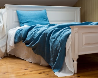 Può includere: Un letto in legno bianco con un piumino e un cuscino in lino blu. Il letto è fatto con un lenzuolo bianco e una coperta blu. La stanza ha un'atmosfera luminosa e ariosa con pavimento in legno e tenda bianca.