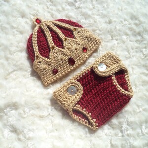 Puede incluir: Un conjunto de gorro y pa&ntilde;al de ganchillo para beb&eacute; en rojo y beige. El gorro tiene forma de corona con adornos dorados y rojos. El pa&ntilde;al tiene dos botones beige.