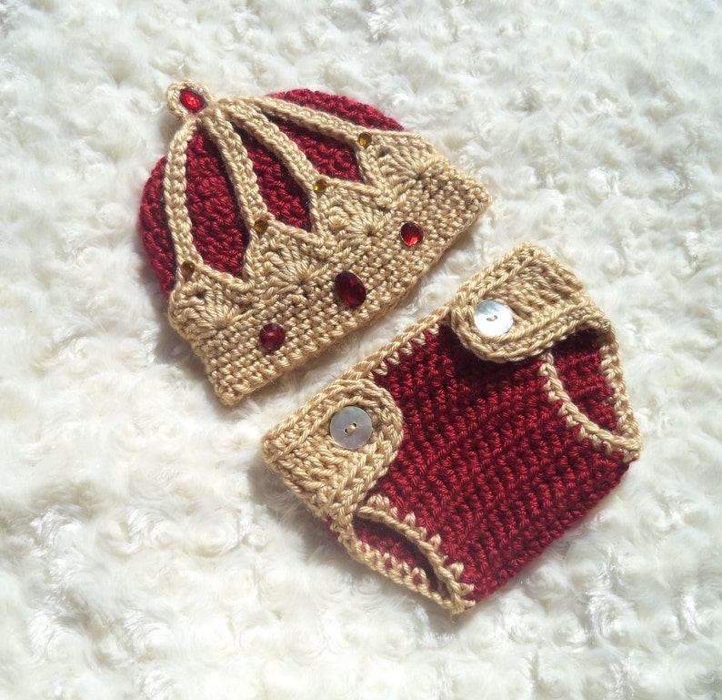Puede incluir: Un conjunto de gorro y pa&ntilde;al de ganchillo para beb&eacute; en rojo y beige. El gorro tiene forma de corona con adornos dorados y rojos. El pa&ntilde;al tiene dos botones beige.