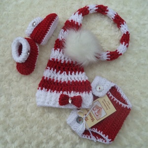 May include: A red and white crocheted baby hat with a white pom-pom, matching booties, and a red and white crocheted diaper cover. The diaper cover has a white bow and a tag that says "Handmade with love".