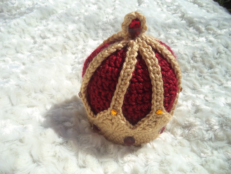 Puede incluir: Una corona de ganchillo de color rojo y dorado. La corona tiene una punta puntiaguda y est&aacute; decorada con peque&ntilde;as gemas brillantes.