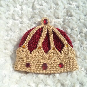 Puede incluir: Un gorro de corona tejido a crochet con la parte superior roja y la inferior dorada. La corona est&aacute; decorada con joyas rojas y doradas.