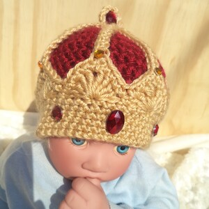 Puede incluir: Un mu&ntilde;eco de beb&eacute; con un gorro de corona de ganchillo con detalles rojos y dorados. El gorro tiene joyas rojas y doradas.