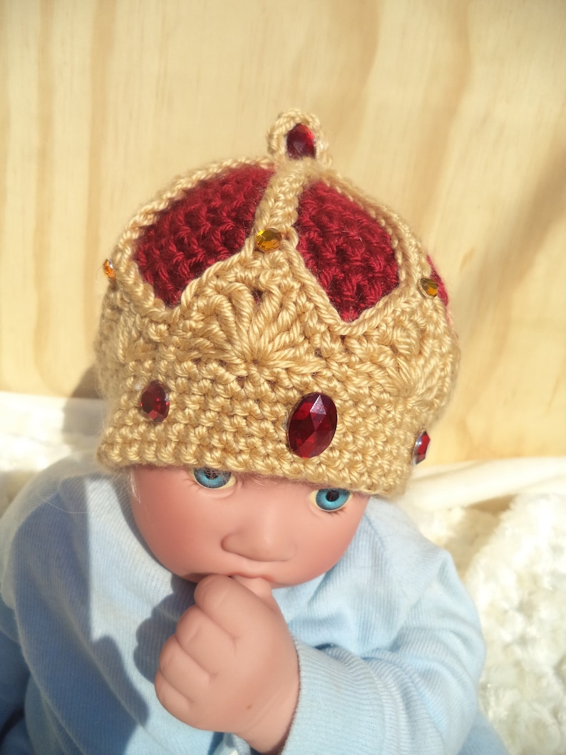 Puede incluir: Un mu&ntilde;eco de beb&eacute; con un gorro de corona de ganchillo con detalles rojos y dorados. El gorro tiene joyas rojas y doradas.