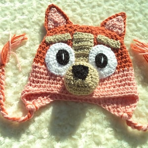 May include: A handmade crochet hat in the shape of a cartoon dog's face. The hat features orange, pink, white, black, and tan yarn. The hat has large white eyes with black pupils, a tan nose, and braided ear ties.