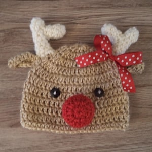 May include: A crocheted reindeer hat with white antlers, a red nose, and a red and white polka dot bow.