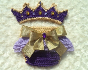 May include: Purple and gold crochet baby outfit with a crown and a bow. The crown is made of yarn and has purple and gold accents. The outfit has a purple crochet skirt with a gold bow and a layer of lace.