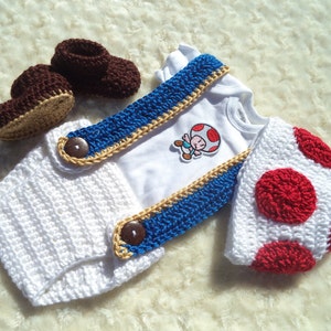 May include: A white baby bodysuit featuring a Toad character patch, paired with white crocheted diaper cover shorts with blue suspenders and brown buttons, a white and red crocheted mushroom cap hat, and a pair of brown crocheted baby booties.