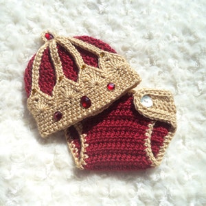 Puede incluir: Un conjunto de beb&eacute; tejido a crochet con un gorro de corona. El gorro es beige con detalles rojos y joyas rojas. El conjunto es rojo con ribetes beige.