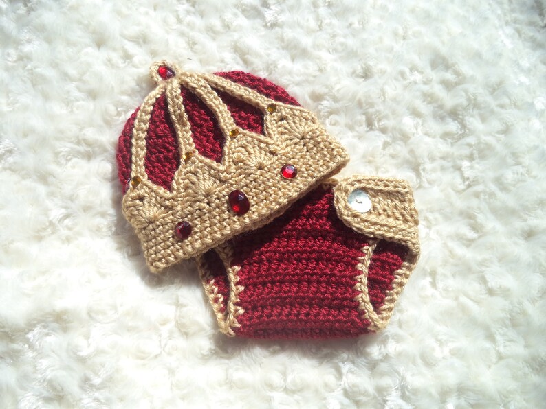 Puede incluir: Un conjunto de beb&eacute; tejido a crochet con un gorro de corona. El gorro es beige con detalles rojos y joyas rojas. El conjunto es rojo con ribetes beige.