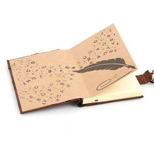 May include: A brown leather-bound journal with a blank page open. The cover is decorated with a pattern of handwritten letters and a feather quill. The journal has a leather strap with a metal clasp.