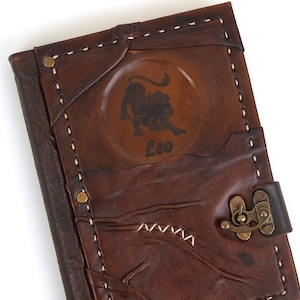 May include: A brown leather journal with a brass clasp and a stitched design. The front cover features a circular embossed design with a lion and the word "Leo".