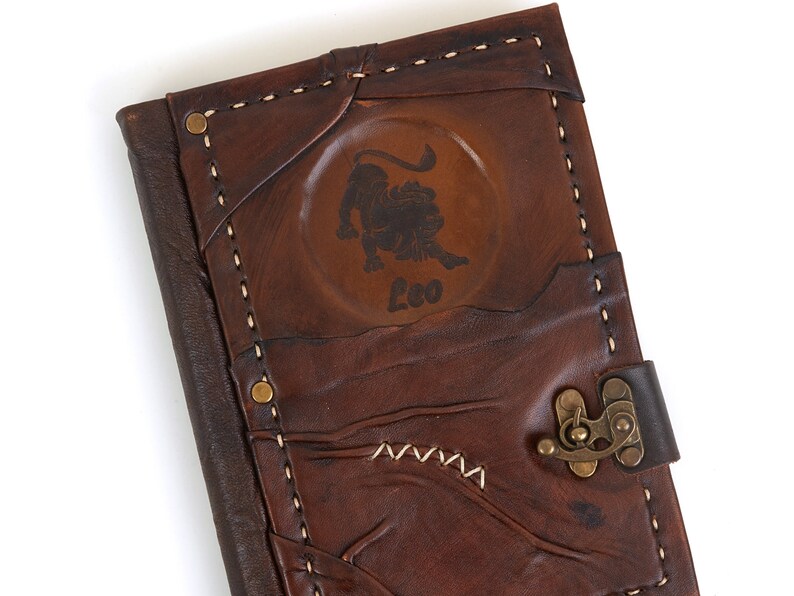 May include: A brown leather journal with a brass clasp and a stitched design. The front cover features a circular embossed design with a lion and the word "Leo".