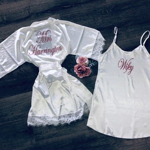 May include: Two white satin robes with lace trim. One robe has "Mrs. Harrington" embroidered on the back in pink. The other robe has "Wifey" embroidered on the front in pink.