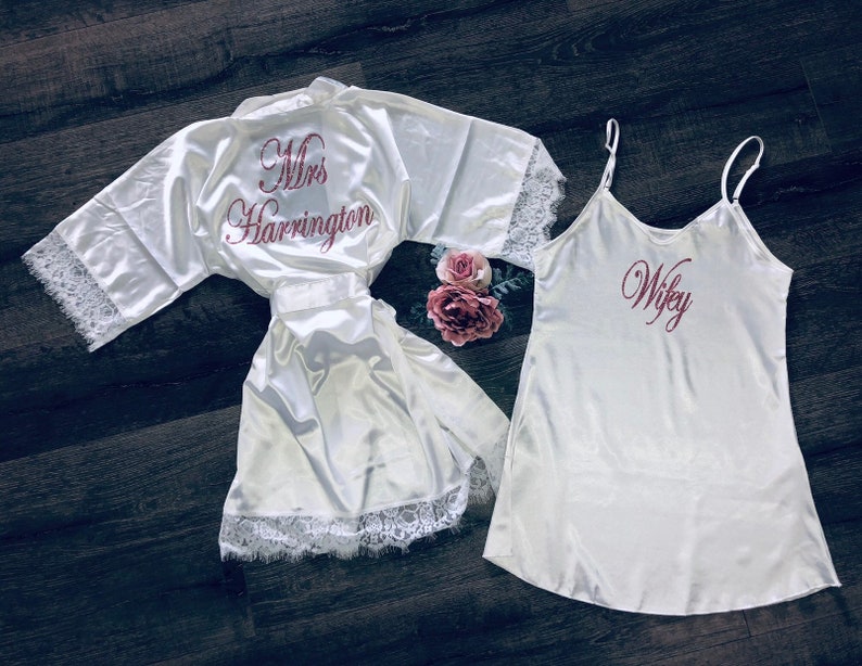 May include: Two white satin robes with lace trim. One robe has "Mrs. Harrington" embroidered on the back in pink. The other robe has "Wifey" embroidered on the front in pink.