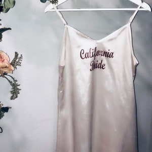 May include: A white satin slip dress with the words "California Bride" embroidered in pink glitter on the front.