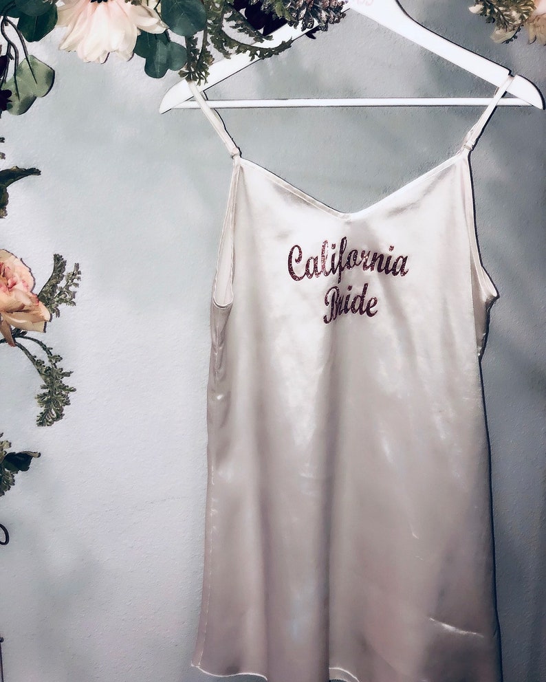 May include: A white satin slip dress with the words "California Bride" embroidered in pink glitter on the front.