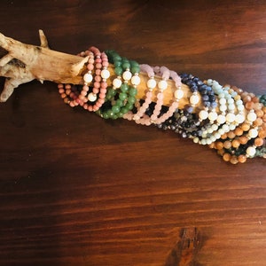 May include: A collection of colourful gemstone bracelets stacked on a piece of driftwood. The bracelets are made with various stones, including green aventurine, rose quartz, amethyst, and lapis lazuli.