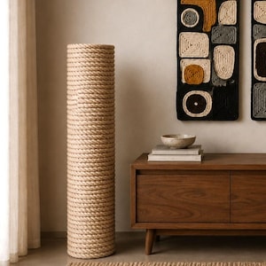 Op de afbeelding: Een hoge, cilindrische krabpaal voor katten, omwikkeld met natuurlijk gekleurd touw. De paal staat naast een donkerhouten kast met een decoratieve wandophanging met geometrische vormen in zwart, beige en geel.