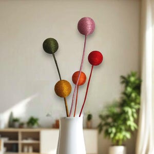 Könnte beinhalten: Eine weiße Vase mit fünf bunten Garnkugeln an Stielen. Die Garnkugeln sind rosa, grün, orange, rot und gelb.