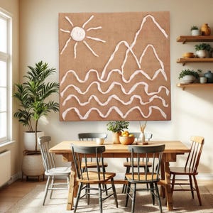 Könnte beinhalten: Ein großes, rechteckiges Leinwandkunstwerk mit braunem Hintergrund zeigt eine weiße Sonne, Berge und wellenförmige Linien. Das Kunstwerk hängt über einem Esstisch aus Holz mit Stühlen.