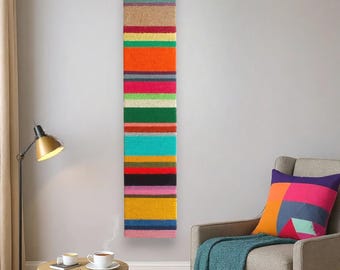 Puede incluir: Una obra de arte mural rectangular vertical con un diseño de rayas coloridas. La obra presenta bandas de varios colores, incluyendo naranja, rosa, verde y azul. Está montada en una pared gris claro, con un sillón gris y un cojín geométrico.
