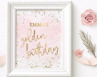 May include: A framed print with the text "EMMA'S golden birthday" in gold script. The background is a soft pink watercolor wash with gold speckles. The white frame has ornate detailing, and a pink rose and greenery are nearby.