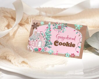 May include: A folded card with a pink background and the words "Gingerbread Cookie" in brown. The card features a gingerbread house illustration with a Christmas tree and snowflake accents. The card is on a white plate.