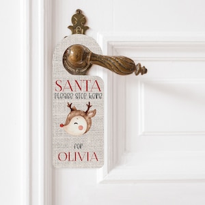 May include: A door hanger with a burlap texture, featuring a watercolor reindeer illustration and the text "SANTA please stop here for OLIVIA" in red and black. The hanger is designed to be placed on a door handle.