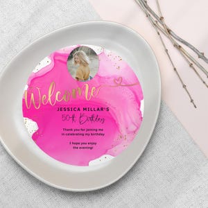 May include: A pink and gold welcome sign with a photo of a person, the text "Welcome", "Jessica Millar's 50th Birthday", and the message "Thank you for joining me in celebrating my birthday. I hope you enjoy the evening!"