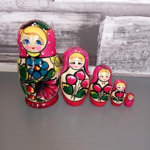 Può includere: Un set di cinque bambole matrioska con disegni floreali dipinti a mano nei toni del rosa, blu, giallo e verde. Ogni bambola in legno presenta un volto sorridente con occhi azzurri e capelli biondi, con la più grande che mostra un fiore blu.