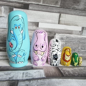 Pode incluir: Um conjunto de seis bonecas russas de madeira pintadas à mão. A boneca maior é azul clara com um elefante. Outras bonecas apresentam um hipopótamo, uma zebra, um leão, um crocodilo e uma pequena boneca amarela.