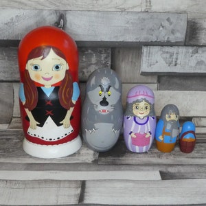 Può includere: Un set di cinque bambole russe annidate, o bambole matrioska, che rappresentano personaggi della fiaba Cappuccetto Rosso. La bambola più grande è Cappuccetto Rosso, che indossa un mantello rosso con cappuccio e un vestito bianco. La bambola successiva è un lupo grigio con un'espressione minacciosa. La terza bambola è una nonna che indossa un vestito viola e un berretto bianco. La quarta bambola è un boscaiolo che indossa una camicia blu e pantaloni marroni. La bambola più piccola è un cestino.